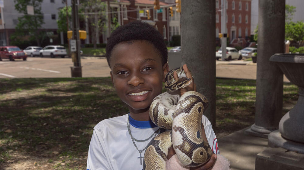 A young smiling Black man holding a snake and standing near a city street.