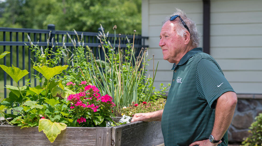 A man, standing beside a wooden table filled with plants, looking into the distance.