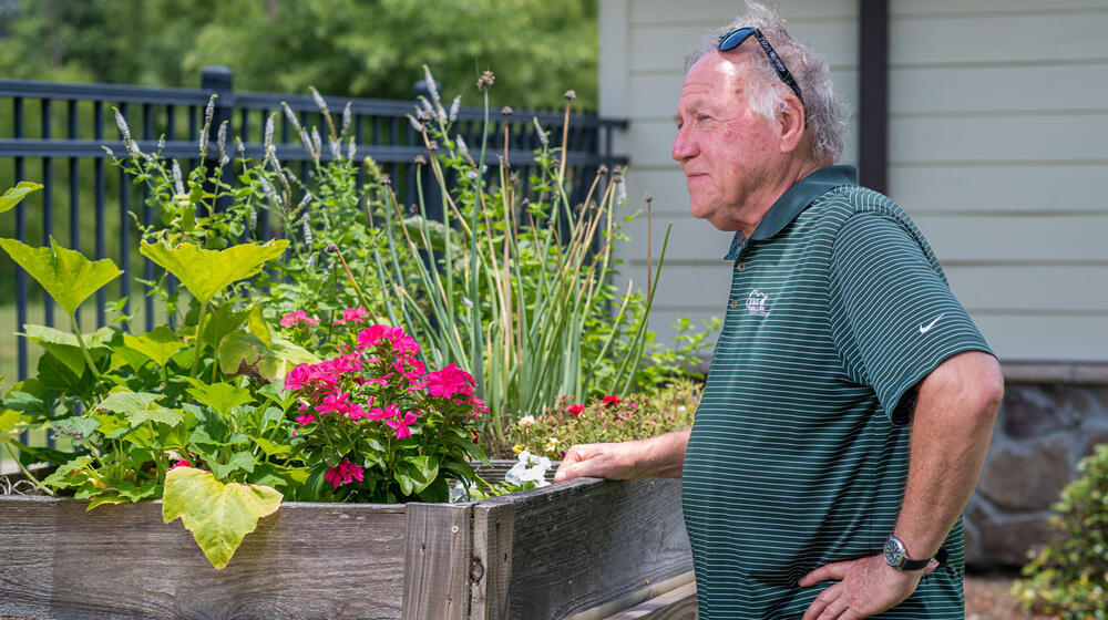 A man, standing beside a wooden table filled with plants, looking into the distance.