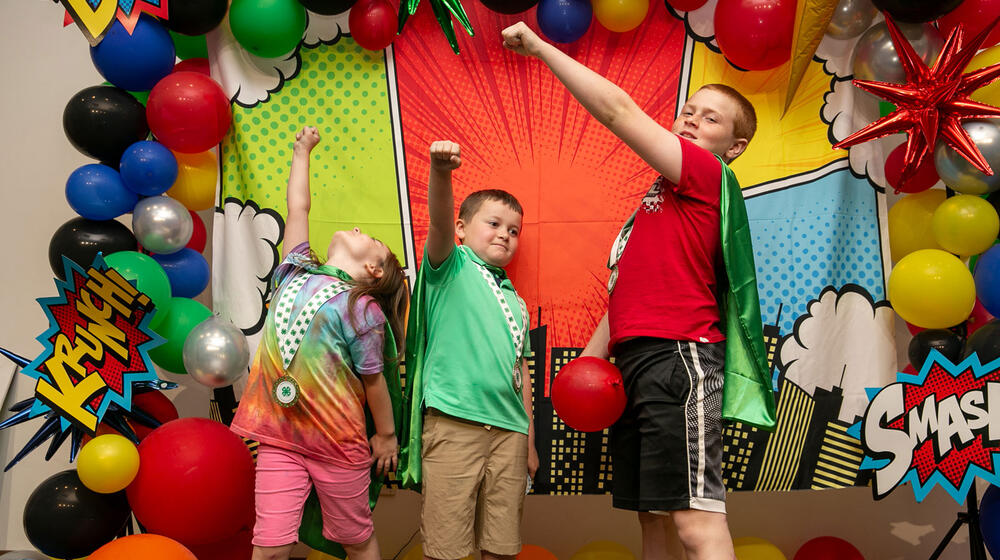 Three children, wearing capes and standing with fists raised.