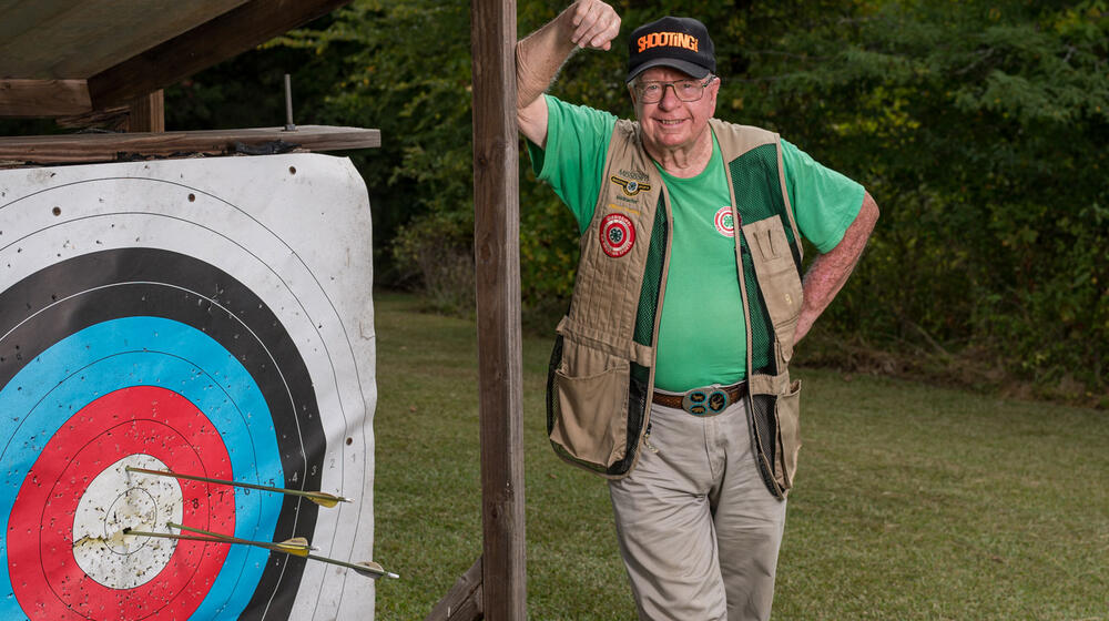 A smiling man wearing a vest and green shirt, standing beside an archery target with three arrows near the bullseye.