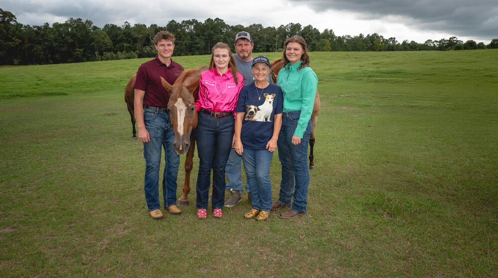 Five people smiling with three horses, all standing on a grassy plain