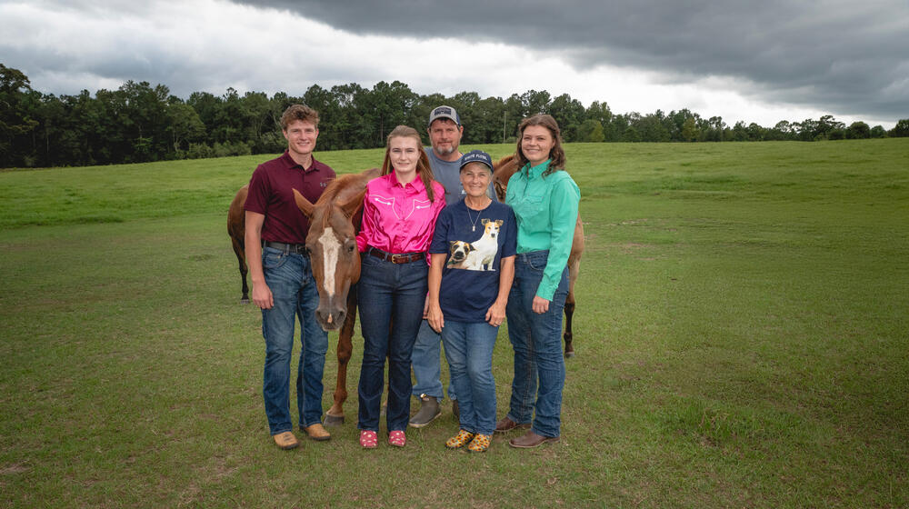 Five people smiling with three horses, all standing on a grassy plain
