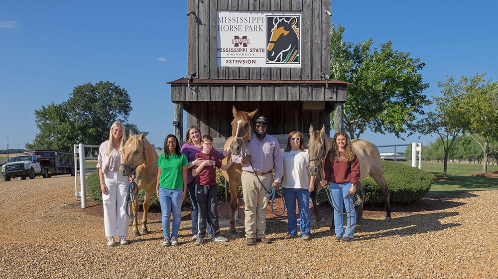 A group of men and women standing in front of a barn and alongside three horses.