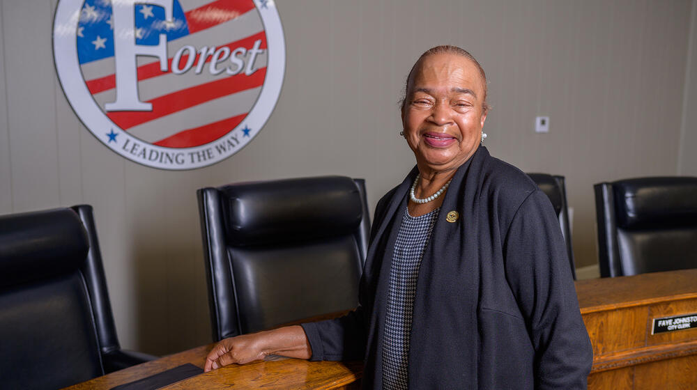 A woman stands smiling in front of the seal for Forest, MS, that lists “Leading the Way” across the bottom.