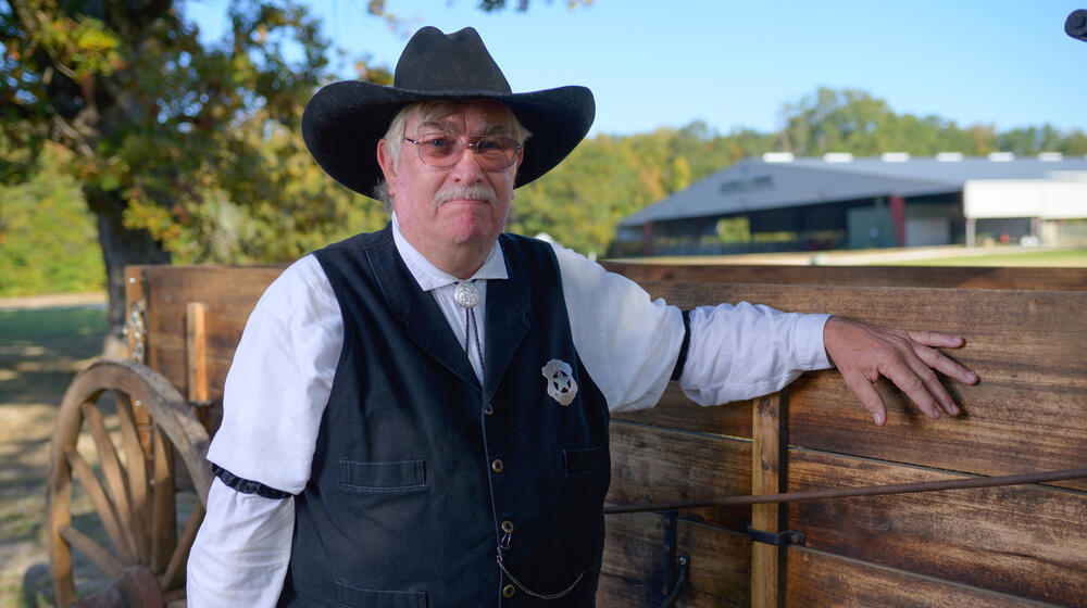 A person with a gray mustache and wearing a black cowboy hat, white polo, and black vest with a sheriff’s star on his chest.