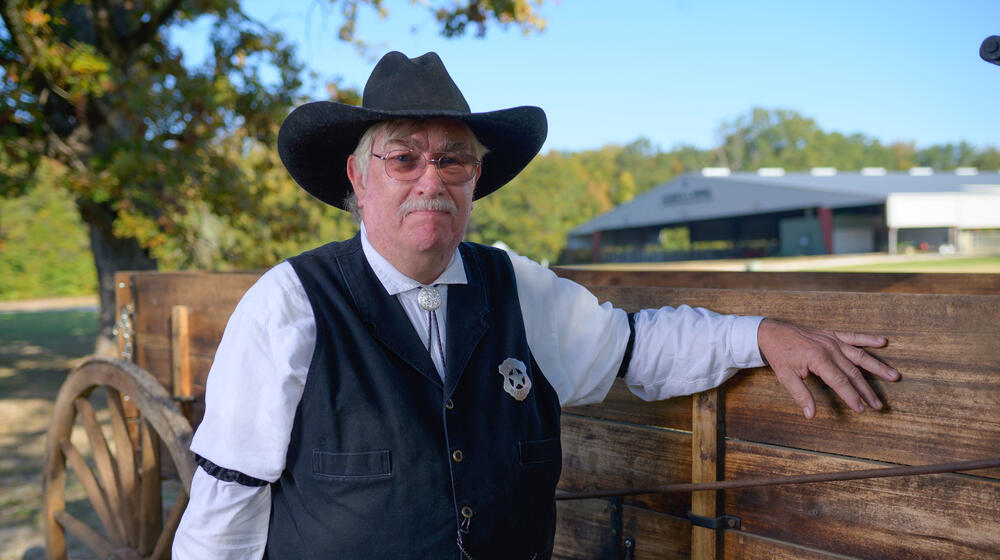 A person with a gray mustache and wearing a black cowboy hat, white polo, and black vest with a sheriff’s star on his chest.