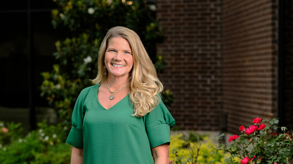 A smiling person wearing green and standing outside in front of roses.