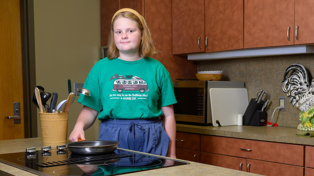A smiling child wearing a green shirt and standing in a kitchen.