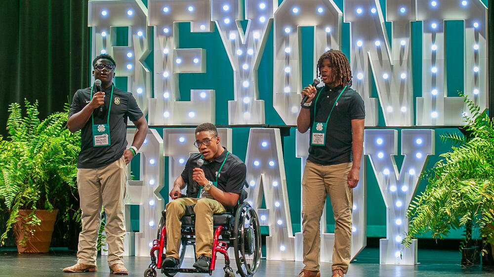 Three teens sing on stage, with the central performer seated in a wheelchair. Large, illuminated letters, arranged to form the words “Beyond Ready,” are in the background.