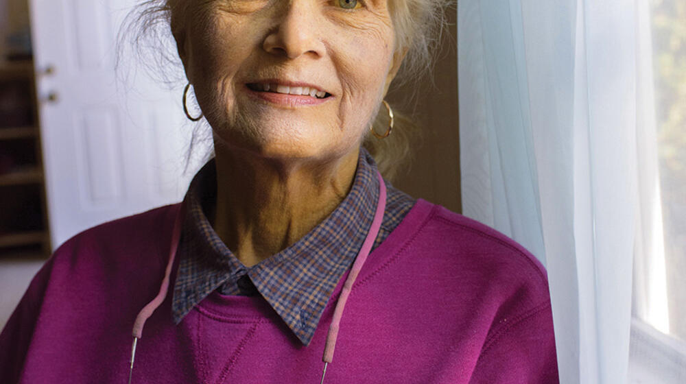 An smiling elderly woman stands next to a window. 