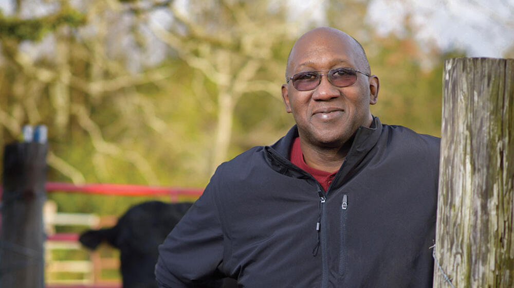 A man wearing all black stands next to a wooden post while a cow peeks out from behind him.