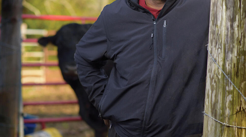 A man wearing all black stands next to a wooden post while a cow peeks out from behind him.