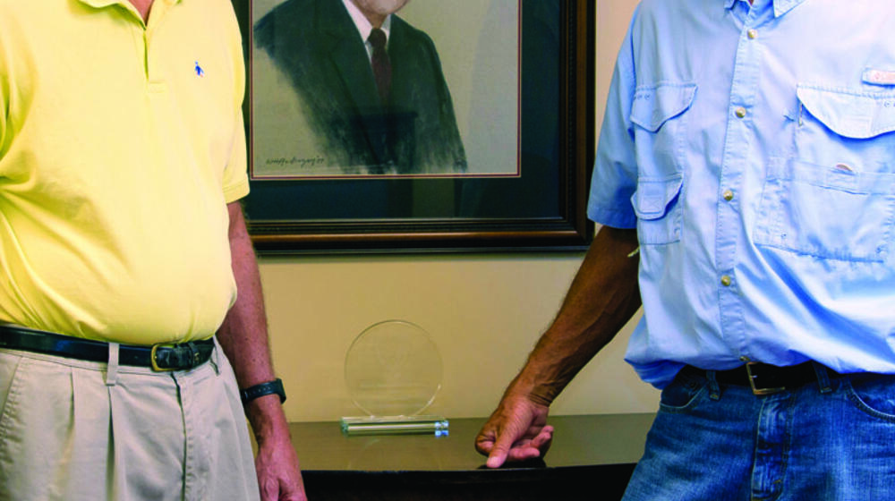 Two men, one in a yellow polo and the other in blue, stand in front of a portrait of a man