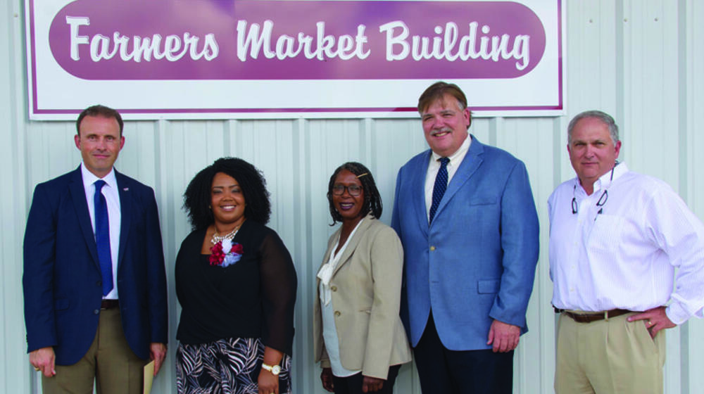 Five people, three men and two women, stand in front of a Farmers Market Building sign