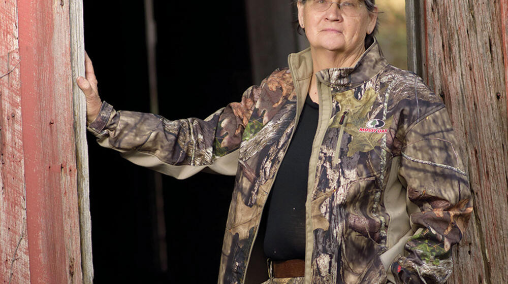 A woman wearing a camouflage jacket and pants rests her hand on the entrance to an old wooden building. 