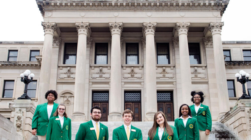 Seven teenagers wearing green blazers and tan slacks form a U standing on steps in front of six gray columns.