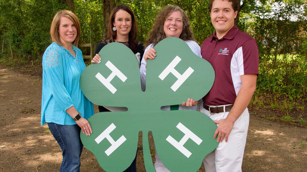 Three women and one man hold a large 4-H clover