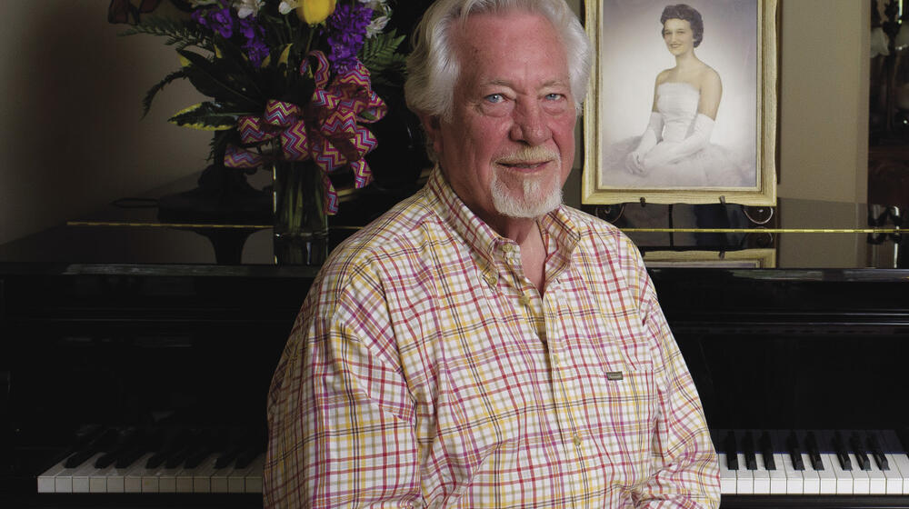 A male retiree in a plaid shirt and khaki slacks sits in front of a piano. On its soundboard is a picture of his late wife in her wedding gown and gloves.