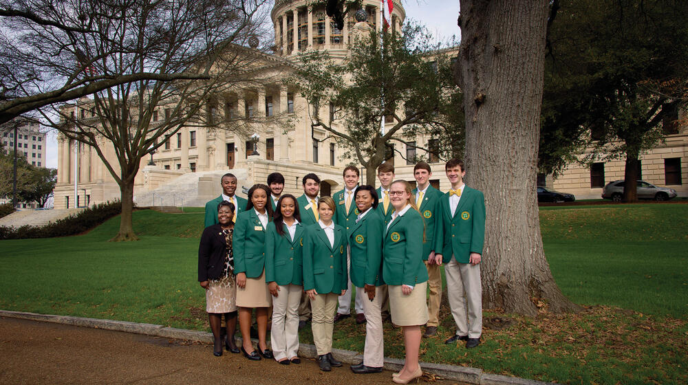 A group of high school girls and boys wearing khaki bottoms and green 4-H blazers stand in a group in front of the state capitol building.