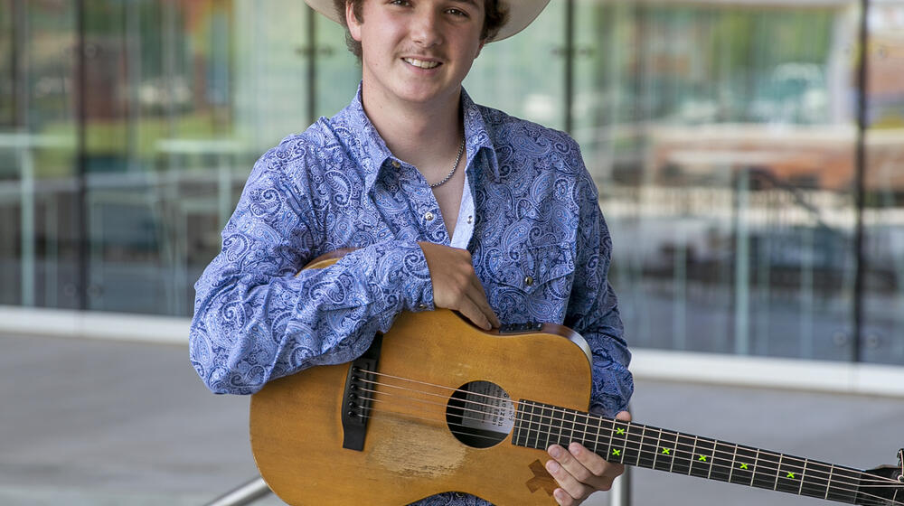 A young man wearing a cowboy hat, paisley collared shirt, and a belt buckle on jeans, holding a guitar and smiling.