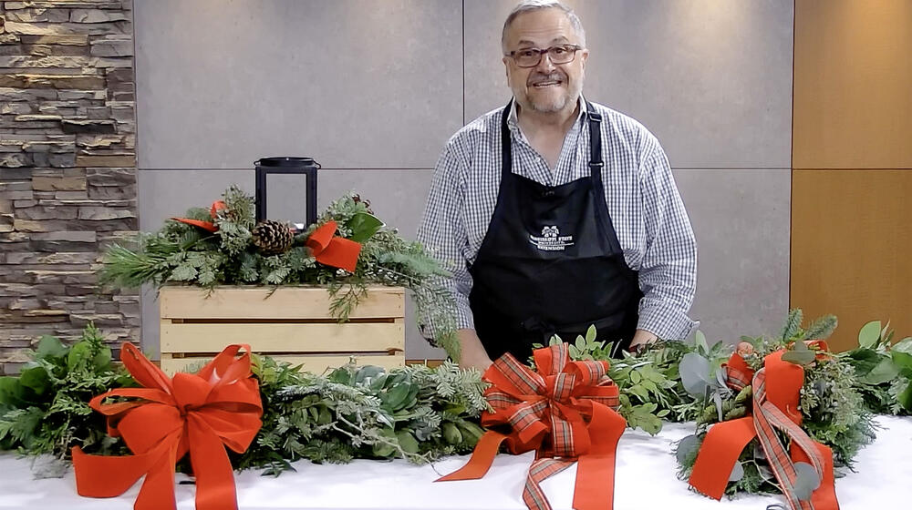 A man, smiling, standing in front of a candle centerpiece and behind a table with bows, evergreen door swag and evergreen garland.