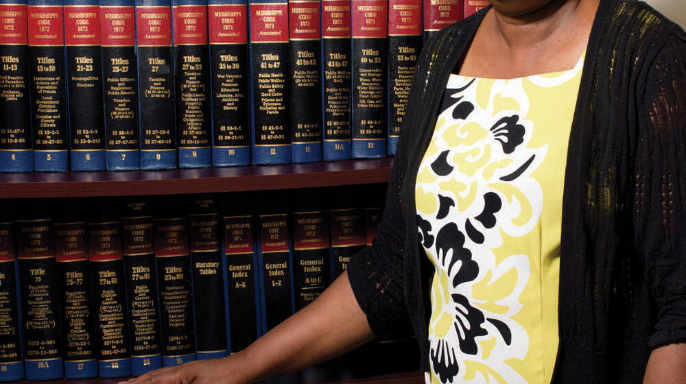 A woman wearing a black, yellow, and white dress stands in front of a bookshelf filled with books.