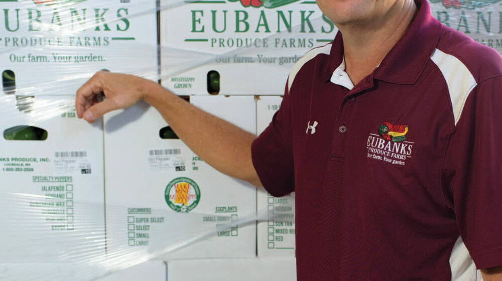 A man wearing a maroon shirt stands in front of a wall of boxes from Eubanks Produce Farm.