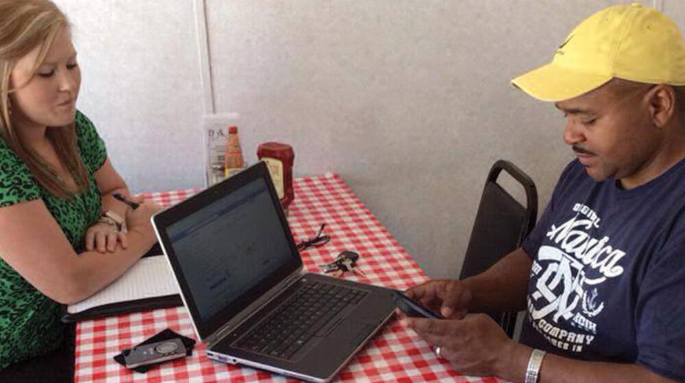 A woman sits on one side of a table, and a man with a laptop sits opposite from her.