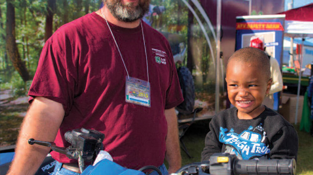 A man stands next to an ATV with a young boy sitting on the seat.
