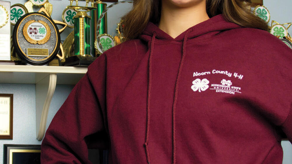 A girl wearing a maroon sweatshirt stands in front of a trophy display with her hands on her hips.