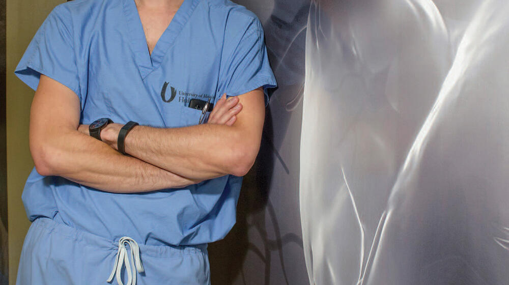 A man in blue scrubs with crossed arms leans against a wall.