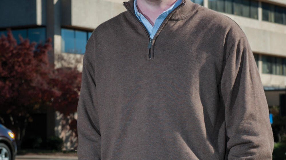 A man with hands in his pockets stands smiling in front of a large concrete building.