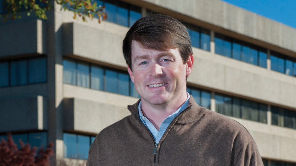 A man with hands in his pockets stands smiling in front of a large concrete building.