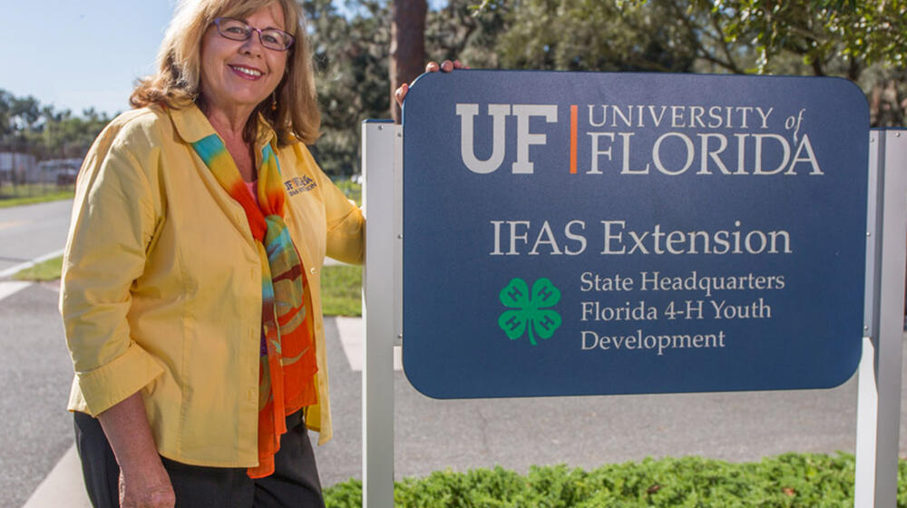 A blonde woman with glasses, wearing a yellow shirt and a motley scarf, stands smiling on a sidewalk in front of trees beside a sign marking “UF University IFAS Extension State Headquarters Florida 4-H Youth Development.”