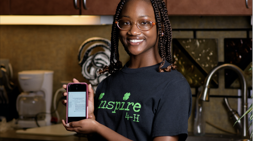 A smiling teenage girl stands behind a table covered in ingredients for a recipe displayed on a phone screen she holds in front of her. 