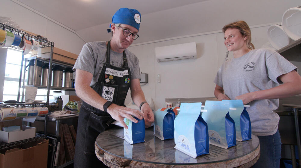 : A man with a blue cap and glasses grabbing a blue bag of coffee for a blonde, smiling woman.