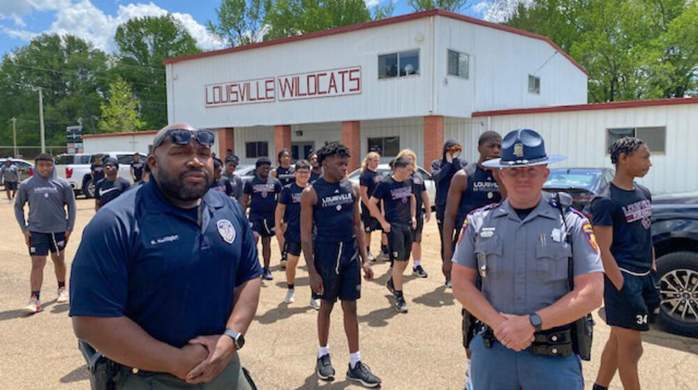 Two officers stand in front of a group of young men spaced apart for workouts in a parking lot.