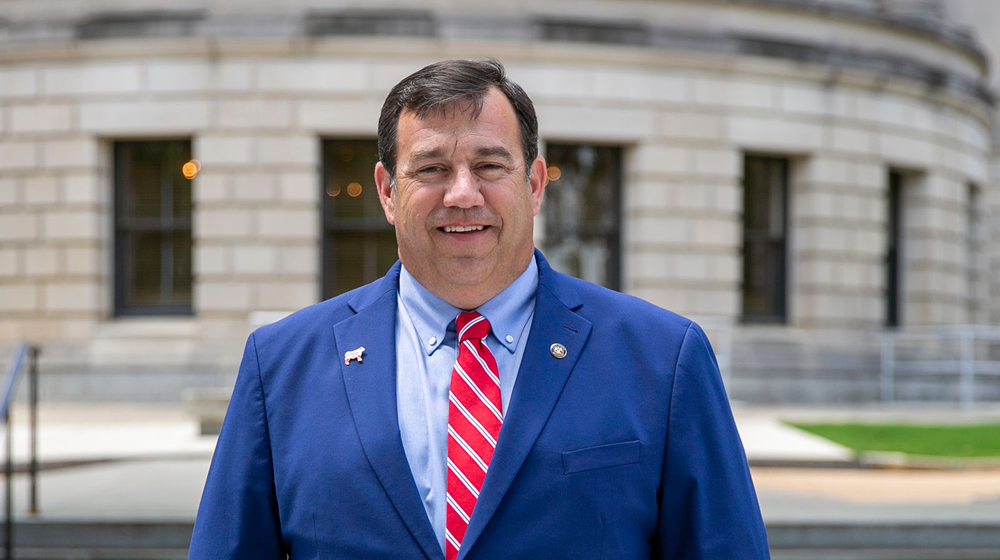 A man, smiling, standing outside and wearing a blue suit with a white-striped red tie.