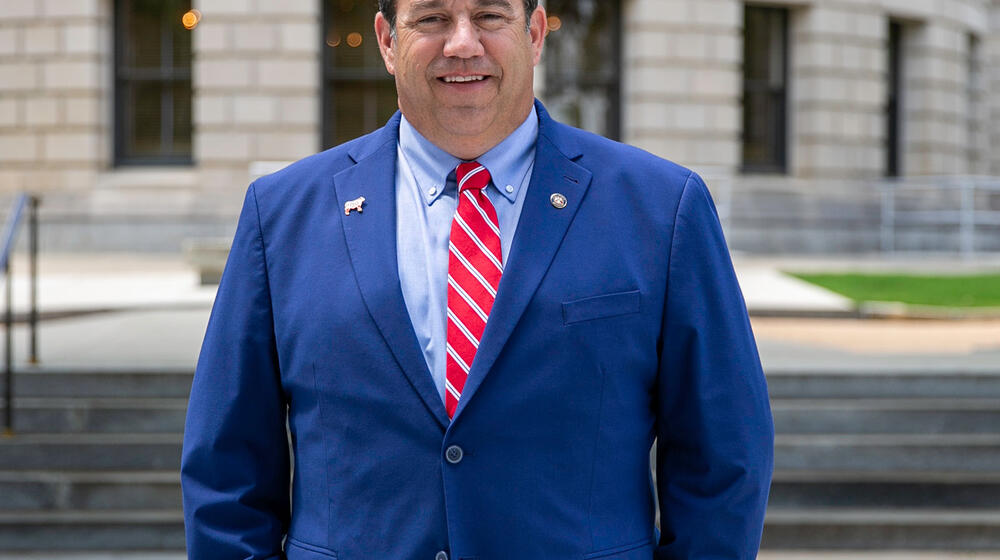 A man, smiling, standing outside and wearing a blue suit with a white-striped red tie.