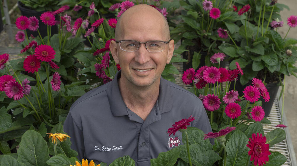 A smiling man with a polo listing his name as “Dr. Eddie Smith” is surrounded by colorful flowers.