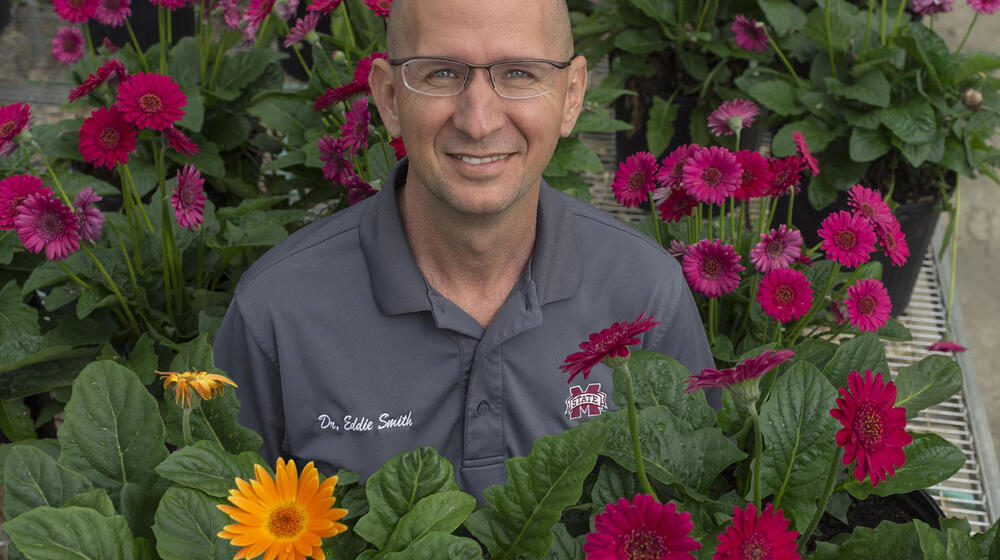 A smiling man with a polo listing his name as “Dr. Eddie Smith” is surrounded by colorful flowers.