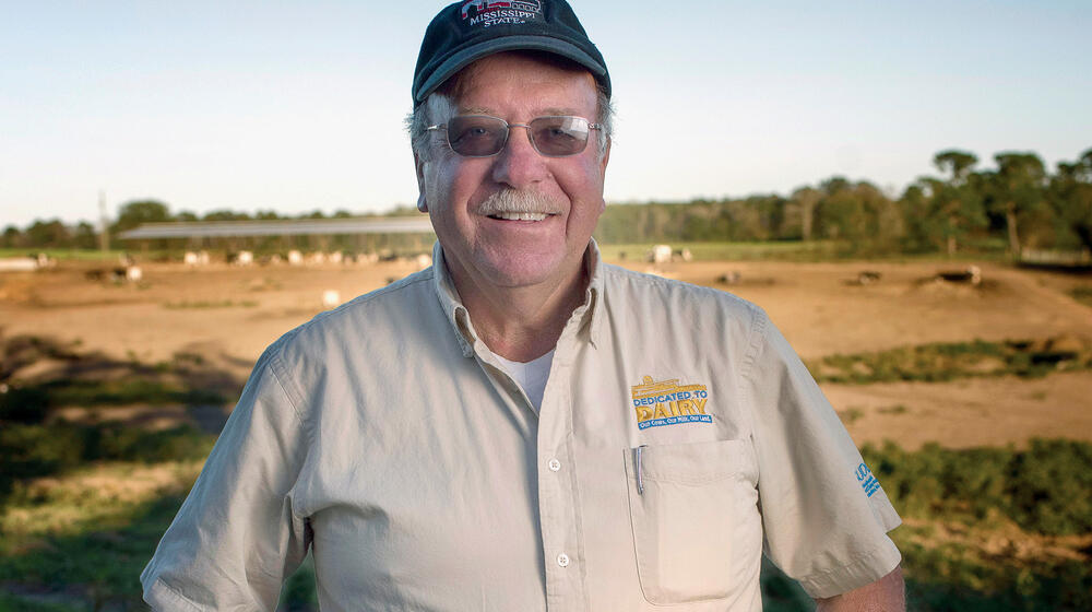 A smiling man with a "dedicated to dairy" logo on his shirt, stands in a pasture.
