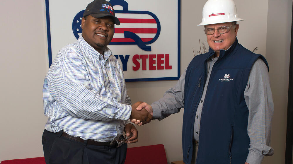 Two men shake hands in front of a Quality Steel sign.