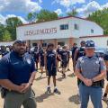 Two officers stand in front of a group of young men spaced apart for workouts in a parking lot.