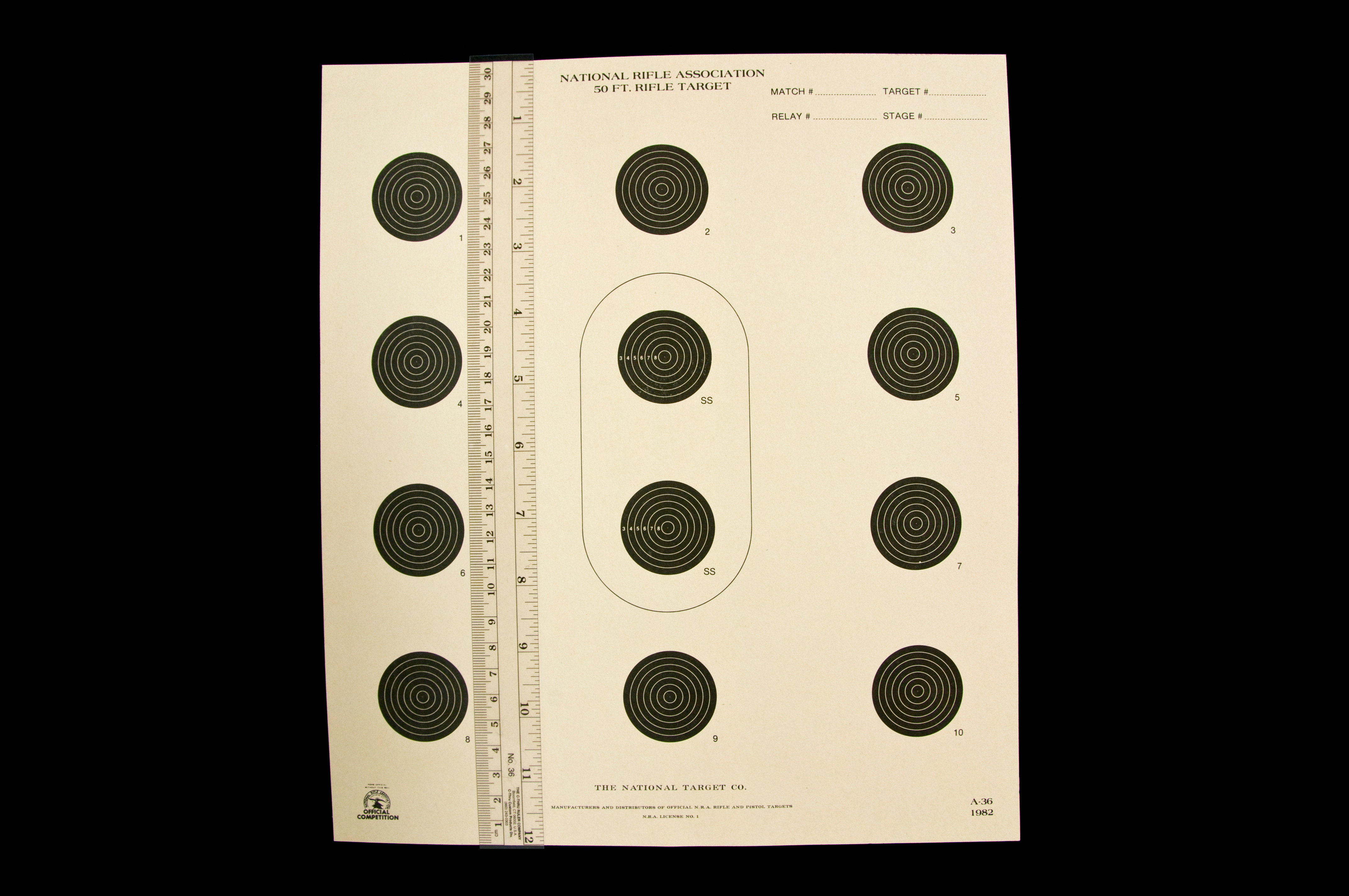 National Rifle Association 50 Ft. Rifle Target with 12 targets and two of them enclosed in an oval with SS labeled on them, all of which have seven black circles getting progressively smaller as they go inward with a ruler lying vertically on the left side of the target.